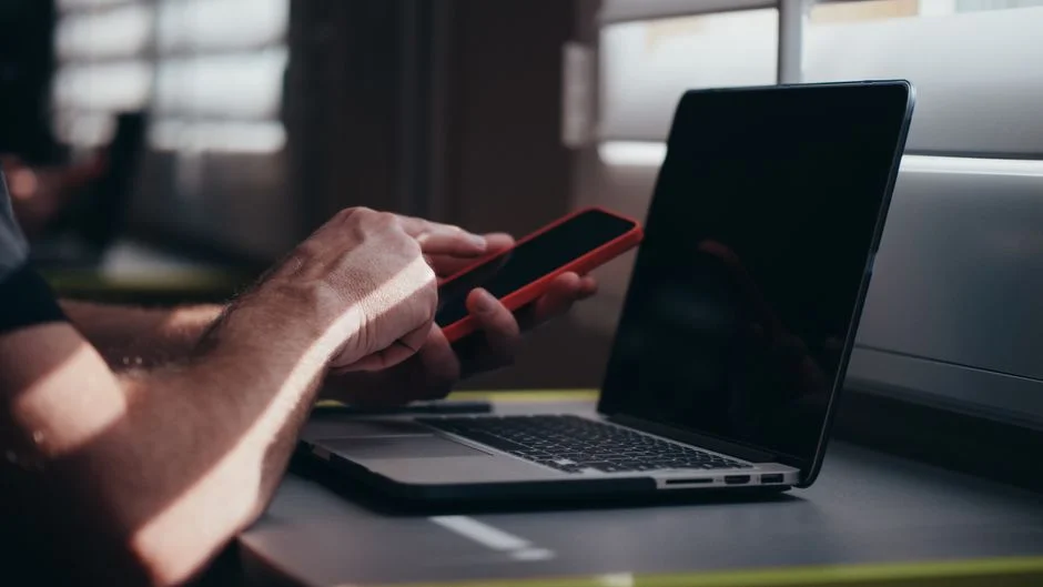 Nahaufnahme einer Hand, die ein Smartphone bedient, im Hintergrund ein aufgeklappter Laptop