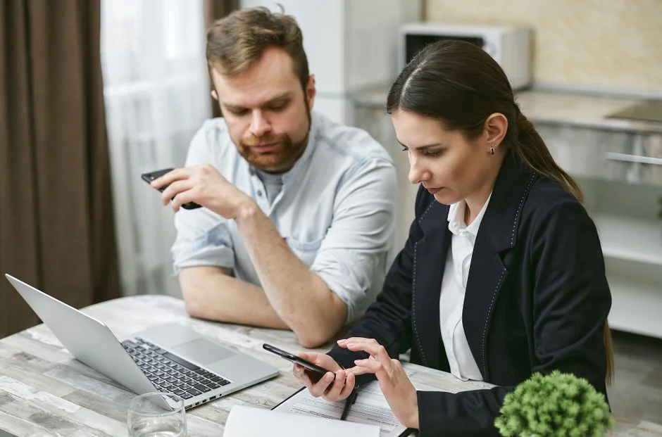 Zwei Personen an einem Besprechungstisch schauen auf ihre Smartphones, daneben ein Laptop